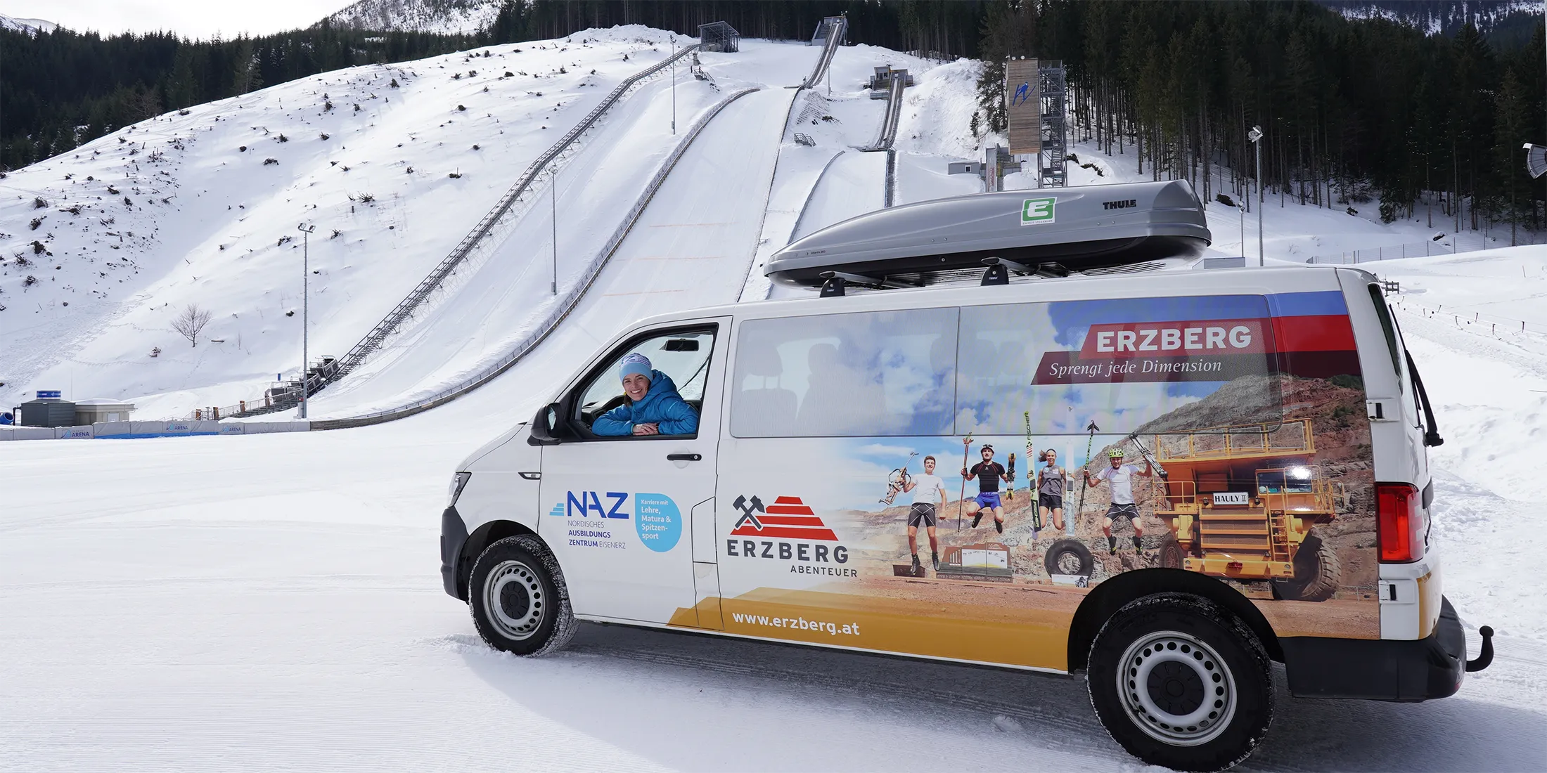 Weißer Minibus mit Erzberg-Werbung und NAZ-Logo, Dame schaut heraus, Schanze im Hintergrund.